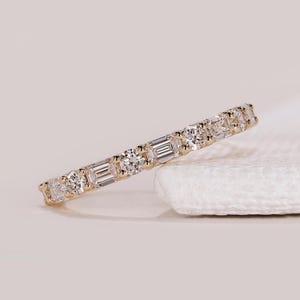 May include: A gold band ring featuring alternating round and emerald-cut clear gemstones. The ring is presented on a white surface against a soft, neutral backdrop. The gemstones are evenly spaced around the band.