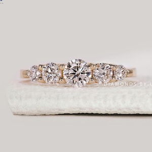 May include: A gold band ring featuring a central, large, round-cut diamond flanked by smaller diamonds on either side. The ring is set on a white surface, showcasing its sparkle and design. The ring is a piece of jewelry.