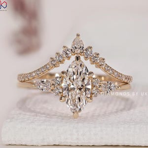 Op de afbeelding: Een gouden ringenset met een centrale diamant in marquise-slijpvorm, geflankeerd door kleinere peervormige en ronde diamanten, gecombineerd met een gebogen band bezet met ronde diamanten. De tekst DIAMONDS BY UK is rechts zichtbaar.