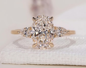 May include: A gold engagement ring featuring a large, oval-cut diamond center stone, flanked by two pear-shaped diamonds. The ring is set on a delicate band, showcasing the brilliance of the diamonds. The ring is displayed on a white surface.