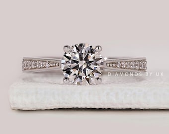 May include: A silver engagement ring with a large, round, clear diamond center stone. The band features smaller diamonds set along the sides. The ring is displayed on a white surface. Text on the image reads "DIAMONDS BY UK".