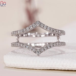 May include: A silver ring with a V-shaped design, encrusted with numerous small, sparkling diamonds. The ring is set against a soft, neutral background, highlighting its intricate details and brilliance. The ring is a jewelry item.