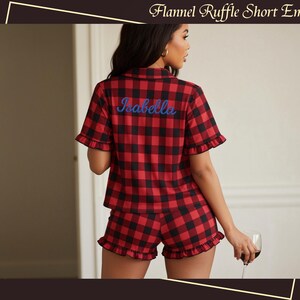 May include: Red and black plaid flannel pajama set with ruffle trim. The short-sleeved top and shorts feature the embroidered name "Isabella" in blue. The set includes a collared shirt and shorts.