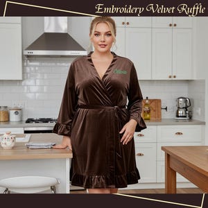 May include: A brown velvet robe with ruffle trim and a matching belt. The robe has long sleeves and a wrap-around design. The name "Olivia" is embroidered in green on the chest. The text "Embroidery Velvet Ruffle Robe" is at the top.