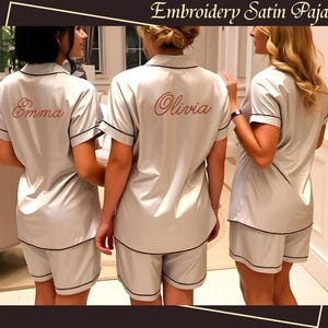 May include: Three women wearing matching light gray satin pajamas with black trim. The pajamas include a short-sleeved top and shorts. The word "Emma" is embroidered on the back of one top, and "Olivia" on another. The image is framed with a brown border and the text "Embroidery Satin Pajamas".