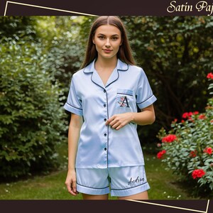 May include: Light blue satin pajama set with black trim. The short-sleeved top has a breast pocket with an embroidered initial and the shorts have a name embroidered on the leg. The words "Satin Pajamas" are in the top right corner.