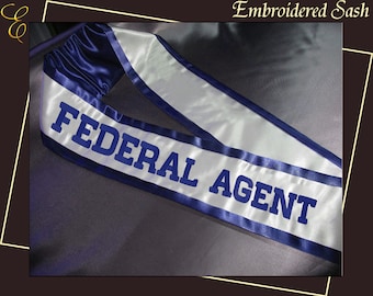 Op de afbeelding: Geborduurde sjerp met de woorden "FEDERAL AGENT" in marineblauwe blokletters. De sjerp is wit met marineblauwe randen en randen. De tekst "Embroidered Sash" staat bovenaan de afbeelding.