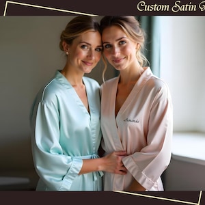 Könnte beinhalten: Zwei Frauen tragen passende Satin-Roben, eine in hellblau und eine in rosa. Die Roben sind mit den Namen "Amanda" und "[Name]" auf der linken Seite der Roben bestickt.