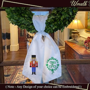 May include: White fabric sash with a green wreath and the letter "K" embroidered on one side and a red and blue nutcracker embroidered on the other side. The sash is hanging from a green boxwood wreath.