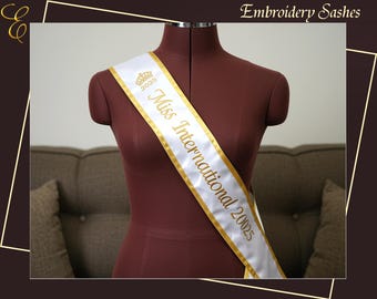 Op de afbeelding: Een witte satijnen sjerp met gouden rand en belettering, gedrapeerd over een mannequin. De sjerp leest "Miss International 20025" in gouden letters, met een gouden kroongrafiek. Een formeel accessoire.