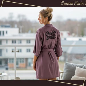 Peut inclure: Robe en satin mauve avec une ceinture assortie, portant l'inscription "Cheerleader" en noir au dos. La robe a des manches trois-quarts. Une personne porte la robe sur un balcon donnant sur un paysage urbain.