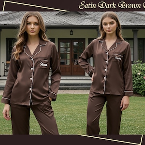 May include: Satin dark brown pajamas set. The set includes a button-down shirt with white trim and a breast pocket, and matching pants. The shirt has the name "Shea" embroidered on the pocket. The pajamas are made of a shiny, dark brown fabric.