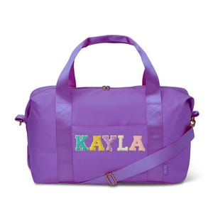 May include: A purple duffel bag with the name "KAYLA" embroidered on the front in colorful letters. The bag has a shoulder strap and two handles. The bag is made of a smooth, durable material.