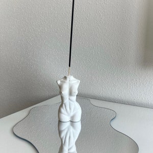 Könnte beinhalten: Ein weißer, minimalistischer Räucherstäbchenhalter in Form eines weiblichen Torsos. Ein schwarzer Räucherstäbchen ist in den Halter eingesetzt. Der Halter spiegelt sich in einer wellenförmigen, spiegelartigen Oberfläche, wodurch ein symmetrisches Bild entsteht.