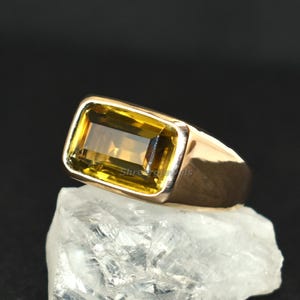 Peut inclure: Une bague en or avec une pierre précieuse jaune rectangulaire. La bague est posée sur une pierre blanche rugueuse sur fond noir. La pierre est de couleur ambre clair et sertie dans une lunette en or.