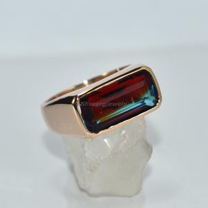 Könnte beinhalten: Ein roségoldener Ring mit einem rechteckigen, mehrfarbigen Edelstein. Der Edelstein zeigt Farbtöne von Rot, Blau und Grün. Der Ring ist auf einer klaren, kristallinen Oberfläche platziert. Der Text "Shreegjewels" ist sichtbar.