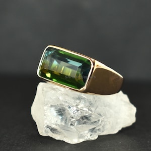 以下が含まれることがあります： 緑と青の色合いの長方形のエメラルドカットの宝石をあしらったゴールドリング。 リングは、暗い背景に対して透明で結晶質の表面に表示され、ジュエリーのデザインを際立たせています。