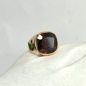 Puede incluir: Un anillo de oro con una gran piedra preciosa de granate rojo oscuro, tallada en forma de cojín y montada en una banda de metal pulido. La piedra tiene una superficie facetada que refleja la luz, y el engaste es liso y redondeado.