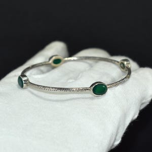May include: A silver bangle bracelet featuring four oval emerald-green gemstones. The bracelet has a textured surface. The bracelet is displayed against a dark background.
