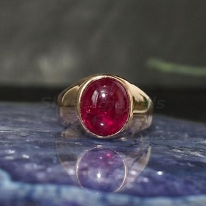 May include: A gold ring with a large, oval, deep red gemstone. The ring's band is polished gold, and the gemstone is set in a bezel. The ring is displayed on a blue surface, reflecting the gemstone.