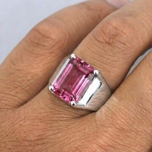 Puede incluir: Un anillo de plata con una gran gema rosa rectangular. El anillo tiene un diseño simple y moderno con un acabado pulido. La gema es de un tono rosa vibrante.