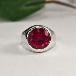 May include: A silver ring with a large, round, deep red gemstone. The gemstone is the focal point, set in a smooth, polished silver band. The ring is displayed on a white surface, with a blurred green leaf and branch in the background.