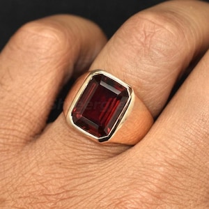 Peut inclure: Une bague en or avec une grosse pierre précieuse rectangulaire rouge foncé. La bague est sertie dans un style simple et classique, mettant en valeur la couleur vibrante de la pierre. La bague est sur le doigt d'une personne.