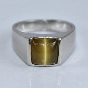 Op de afbeelding: Een zilverkleurige ring met een gladde, vierkant geslepen, goudbruine kattenoog-edelsteen in het midden. De ringband is breed en gepolijst, met een solide metalen constructie die iets taps toeloopt naar de achterkant van de ring.