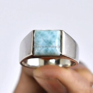 May include: A silver-toned ring featuring a square-cut, light blue, marbled stone set in the center. The metal band is smooth and polished, with the text shreejewels visible across the surface of the blue stone in a light, subtle font.