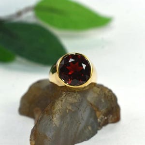 Puede incluir: Un anillo de oro con una gran piedra preciosa de granate rojo oscuro, redonda y facetada, engastada en una banda de oro lisa y pulida. El anillo está colocado sobre una piedra gris translúcida y texturizada, resaltando su color y brillo.