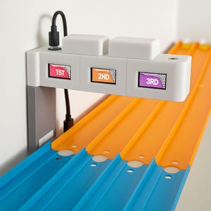 Op de afbeelding: Een speelgoed racebaan met drie banen in blauw en oranje. Een witte finishlijn toont "1ST", "2ND" en "3RD" met geblokte vlaggen. Een zwarte kabel en een USB-connector zijn bevestigd.