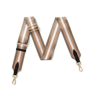 May include: A striped bag strap in shades of brown, white, and tan. The strap features gold-coloured metal hardware, including a buckle and clasps. Black leather accents are visible near the clasps.