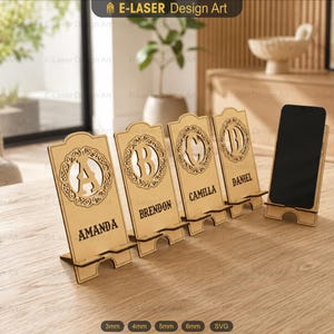 Könnte beinhalten: Vier helle Handyhalterungen aus Holz mit lasergeschnittenen Motiven. Jeder Ständer zeigt ein kreisförmiges Monogramm mit einem Buchstaben und einem darunter eingravierten Namen: AMANDA, BRENDON, CAMILLA und DANIEL. Ein schwarzes Smartphone.