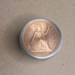 May include: A copper-coloured coin with a silver rim. The coin features a woman sitting on a throne with a trident in her hand. The text "ONE PENNY" is inscribed around the woman and the year "1967" is inscribed below her.