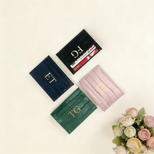 Könnte beinhalten: Vier Leder-Kartenetuis in Marineblau, Schwarz, Grün und Rosa, jeweils mit goldenen Initialen geprägt. Die Kartenetuis haben eine Krokodilleder-Textur. Ein schwarzes Kartenetui enthält Kreditkarten. Ein Blumenstrauß befindet sich in der unteren rechten Ecke.