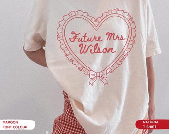 May include: A natural-colored t-shirt featuring a maroon graphic of a heart with a ruffled border and a small bow at the bottom. The text "Future Mrs Wilson" is printed in a cursive script inside the heart. The shirt is worn with red and white checkered shorts.