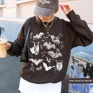 May include: A dark chocolate brown crewneck sweatshirt featuring a white graphic print of various bat species and botanical elements. The long-sleeved pullover has a relaxed fit and is worn with a grey baseball cap and a black shoulder bag.
