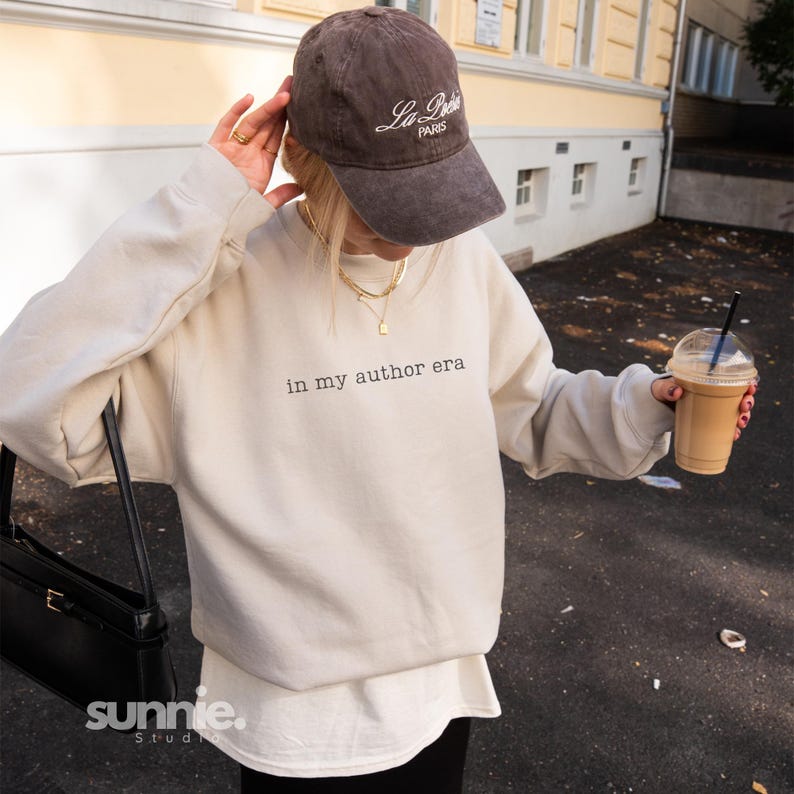 May include: A beige crewneck sweatshirt featuring the black text 'in my author era' on the chest. The outfit includes a dark brown baseball cap with 'La Po&eacute;sie PARIS' embroidered in white, a gold layered necklace, and a black shoulder bag.