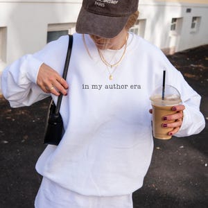 May include: A white crewneck sweatshirt featuring the black text 'in my author era' printed on the chest. The garment has long sleeves and a relaxed fit. It is paired with a dark baseball cap, layered gold necklaces, and a black shoulder bag.