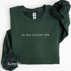 May include: A dark forest green crewneck sweatshirt featuring the white embroidered text "in my author era" centered on the chest. The garment has long sleeves with ribbed cuffs and a ribbed neckline, with a Gildan brand label visible inside.