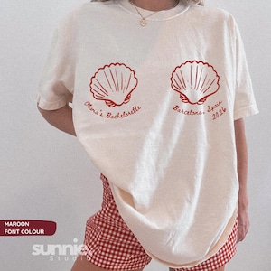 Op de afbeelding: Een off-white t-shirt met korte mouwen en twee rode schelpillustraties. De tekst onder de linker schelp luidt Olivia's Bachelorette en onder de rechter schelp Barcelona, Spain 2026. Het t-shirt wordt gedragen met een rood-witte gingham short.