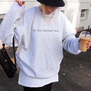 May include: A light gray crewneck sweatshirt featuring the black text "in my author era" printed on the chest. The garment has long sleeves and a relaxed fit, paired with a black bucket hat, layered gold necklaces, and a black shoulder bag.