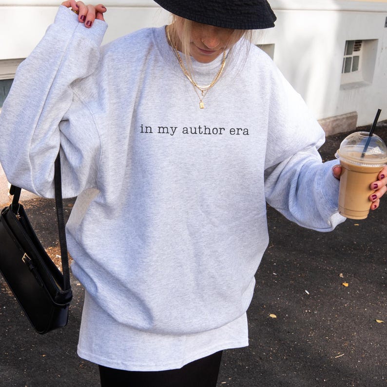 May include: A light gray crewneck sweatshirt featuring the black text "in my author era" printed on the chest. The garment has long sleeves and a relaxed fit, paired with a black bucket hat, layered gold necklaces, and a black shoulder bag.