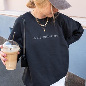 May include: A black crewneck sweatshirt featuring the white text "in my author era" printed across the chest. The garment has long sleeves and a classic fit, providing a simple and clean look for casual wear in various settings and daily activities.