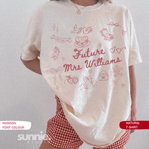 Puede incluir: Una camiseta de color natural con gráficos de líneas en color granate y el texto "Future Mrs Williams". Las ilustraciones incluyen un pastel, un joyero, copas de champán, corazones, lazos, flores y una pancarta con la palabra "BRIDE".