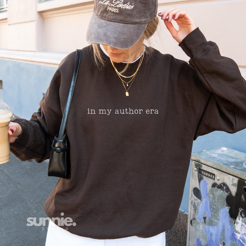 May include: A dark brown crewneck sweatshirt featuring the white text 'in my author era' printed on the chest. The garment is paired with a grey baseball cap with 'La Po&eacute;sie PARIS' text, layered gold necklaces, and a black shoulder bag.