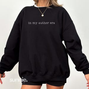 May include: A black crewneck sweatshirt featuring the white text "in my author era" printed across the chest. The garment has long sleeves, a ribbed neckline, and a relaxed fit. A gold pendant necklace is worn around the neck area.