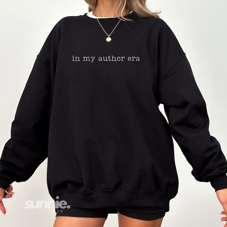 May include: A black crewneck sweatshirt featuring the white text "in my author era" printed across the chest. The garment has long sleeves, a ribbed neckline, and a relaxed fit. A gold pendant necklace is worn around the neck area.