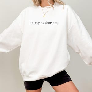 May include: A white crewneck sweatshirt featuring the black text "in my author era" printed on the chest. The garment has long sleeves and a ribbed neckline, cuffs, and hem. A gold-toned chain necklace with a small circular pendant is visible.