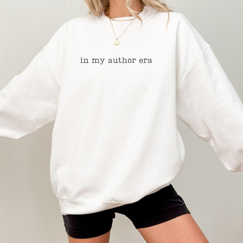 May include: A white crewneck sweatshirt featuring the black text "in my author era" printed on the chest. The garment has long sleeves and a ribbed neckline, cuffs, and hem. A gold-toned chain necklace with a small circular pendant is visible.